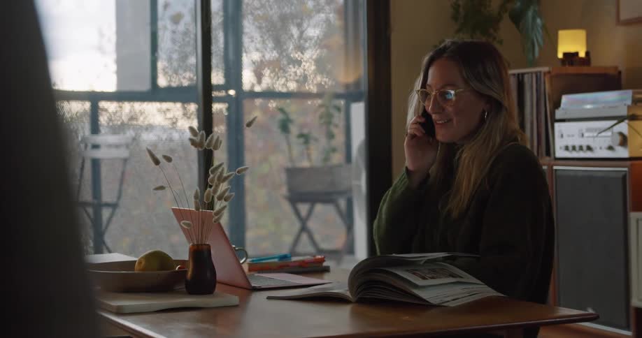 A woman sits at a table talking on the phone with a laptop and a book in front of her