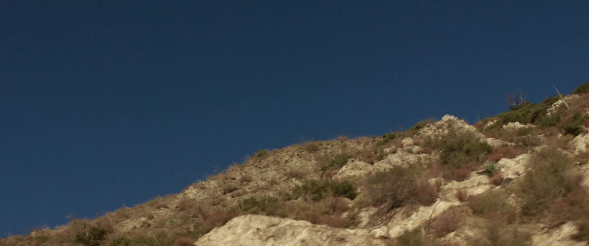Rocky mountain side with harsh plants and a few rough looking shrubs in front of a deep blue sky