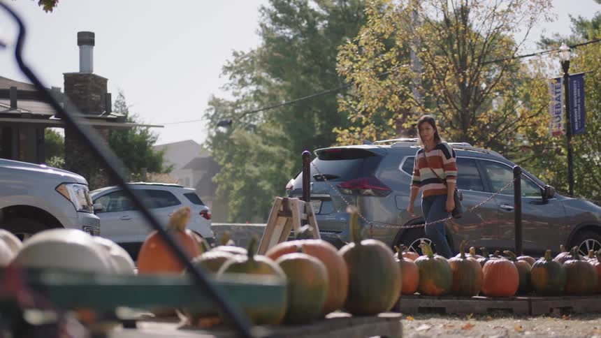 A woman walks on a sidewalk toward a farmers market with a large selection of pumpkins