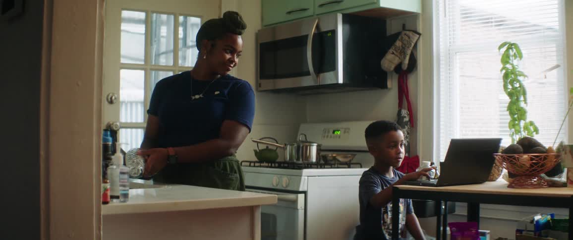 Mother with earring and a headband does the dishes while son looks at laptop in the kitchen
