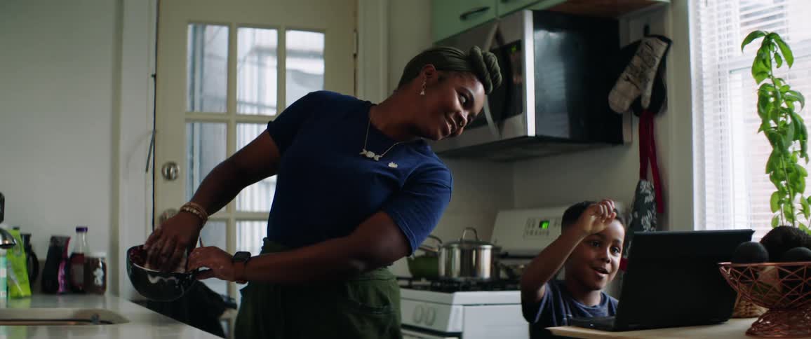 Mother with earrings and a headband does the dishes while son looks at a laptop in the kitchen