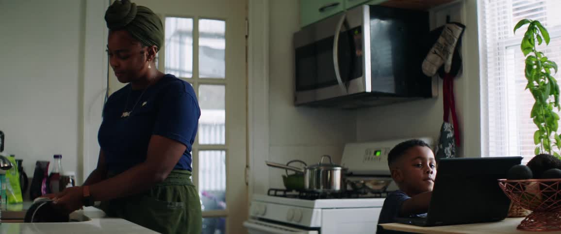 Mother with earrings and a headband does the dishes while son looks at laptop in the kitchen