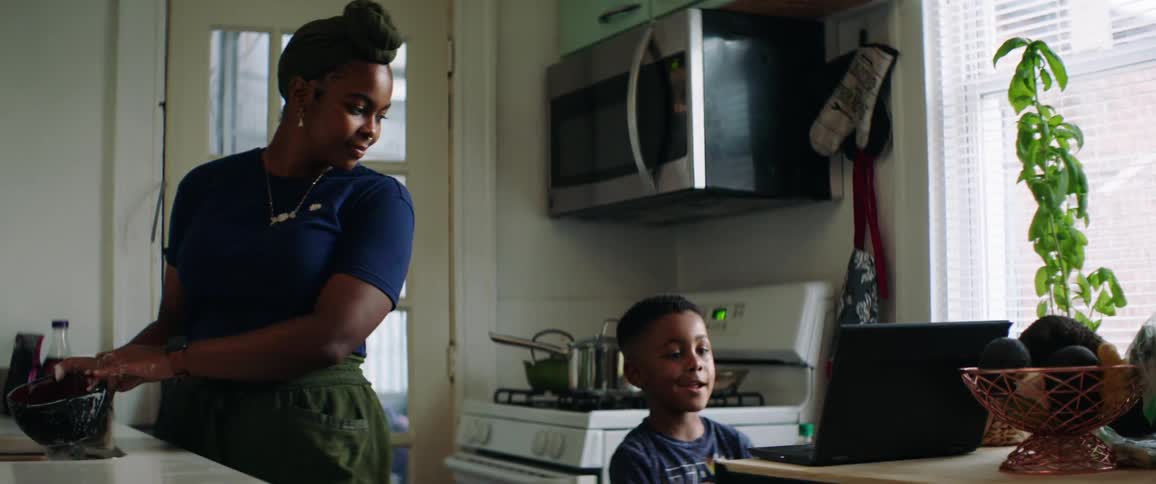 A woman washing a bowl in a kitchen sink before pointing something out on the screen of a nearby laptop for a little boy