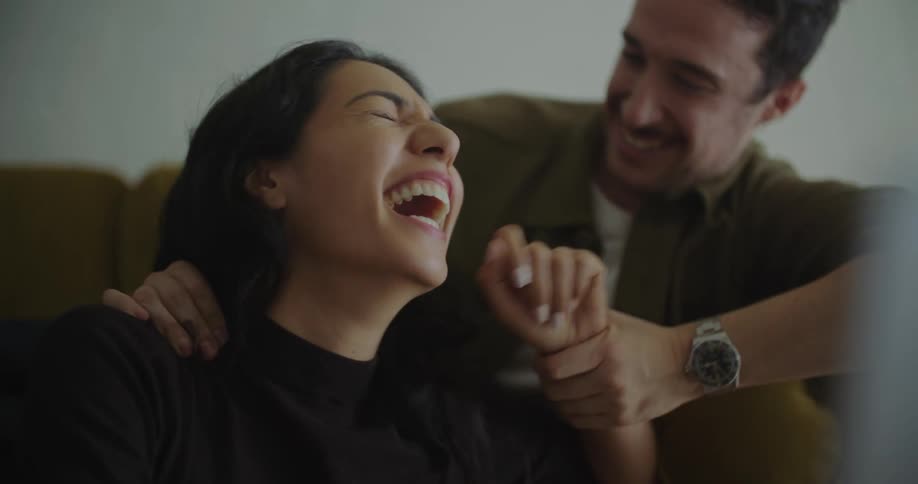 Young man and woman sitting on the couch together laughing and smiling while watching something on laptop