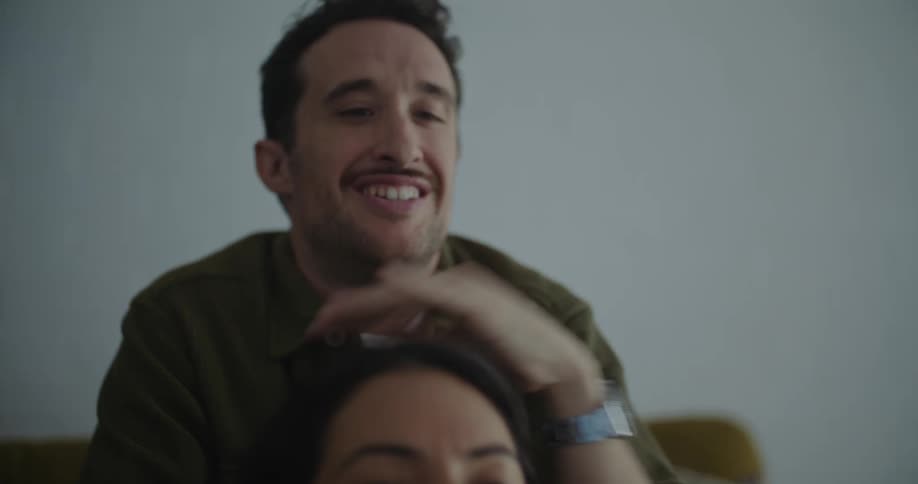 A woman is sitting on the floor holding a remote controller with a frustrated look on her face and the man sitting behind her puts his hand on her head and takes it from her and they laugh