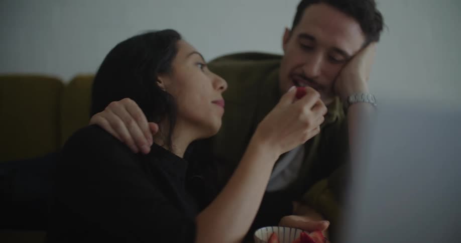 A woman feeds strawberries to her partner while using a laptop together