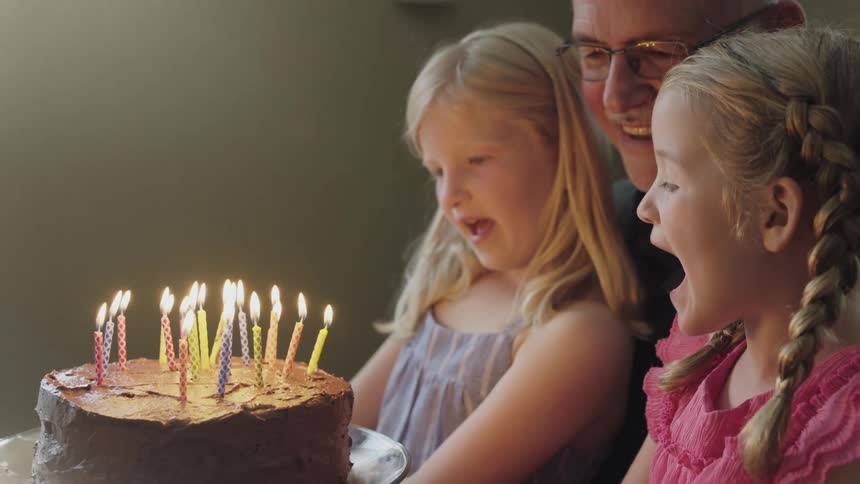 Grandfather sits close to granddaughters next to birthday cake with lit candles while girls sing