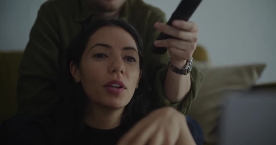 A woman is sitting on the floor in front of a table looking at a laptop computer as a man is sitting on the couch behind her holding a remote controller in his hand