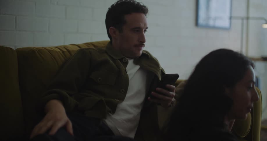 A woman is sitting on the floor in front of a table with a glass of juice and a plate of food looking at a laptop computer as a man is sitting on the couch behind her texting on his cell phone