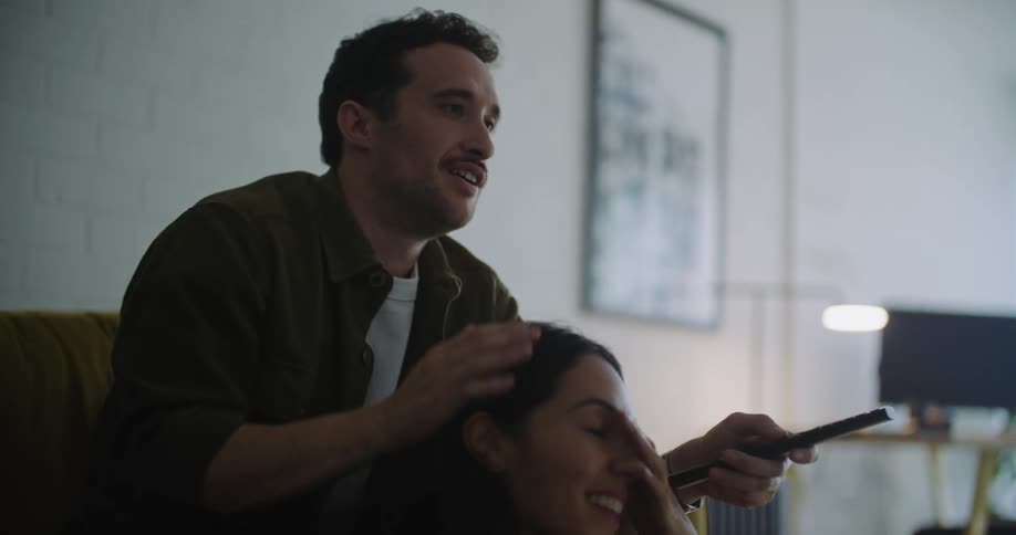 Young couple sitting on couch together watching tv laughing and smiling he strokes her hair with one hand and remote control in the other