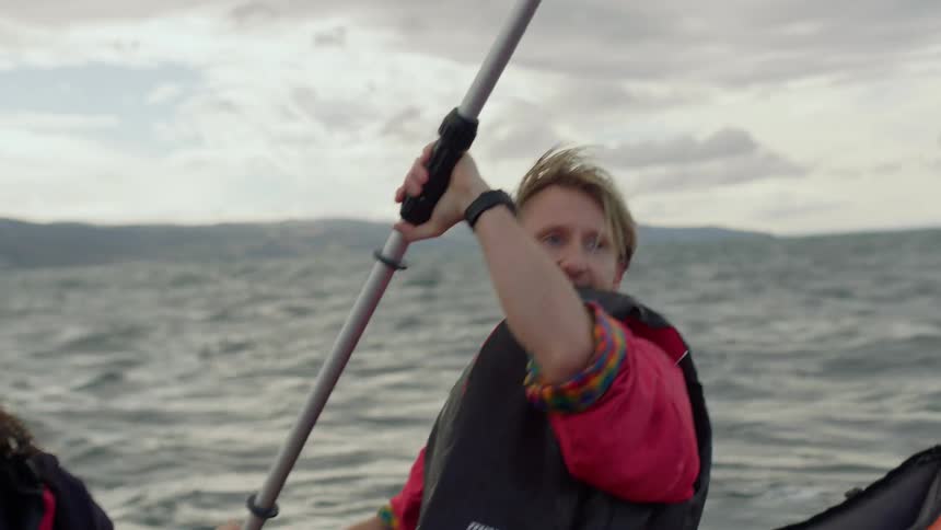 A man is looking around as he rows an inflatable boat across rough water as a woman sits in front of him