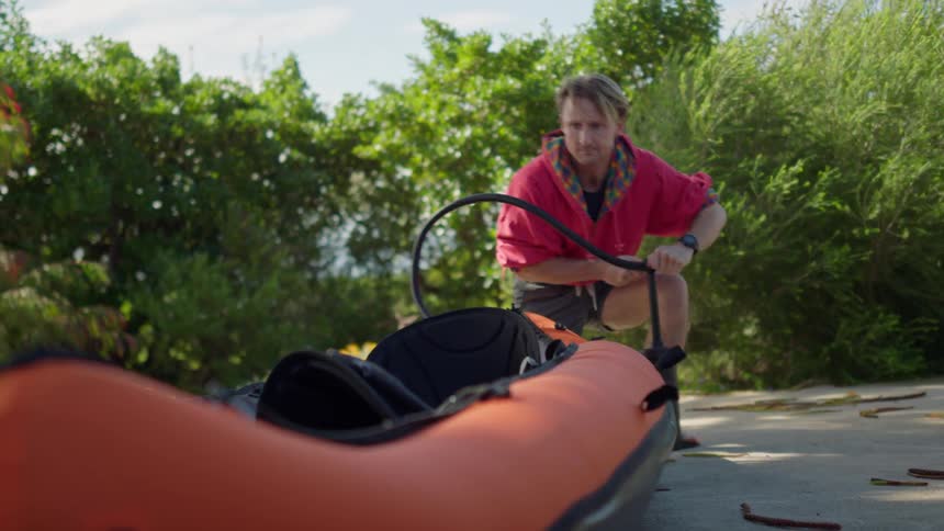 A man is standing outside pumping up an inflatable boat as the sun shines upon the trees as they move in the breeze