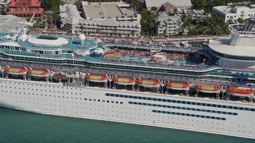 Aerial footage of a large Royal Caribbean cruise ship approaching and flying by while docked at Key West Florida during a sunny daytime with multiple leisure activities visible on board