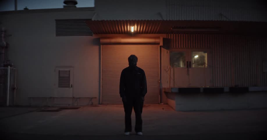 Silhouette of a person standing outside of a building at night with their hands in their pockets as a light shines behind them