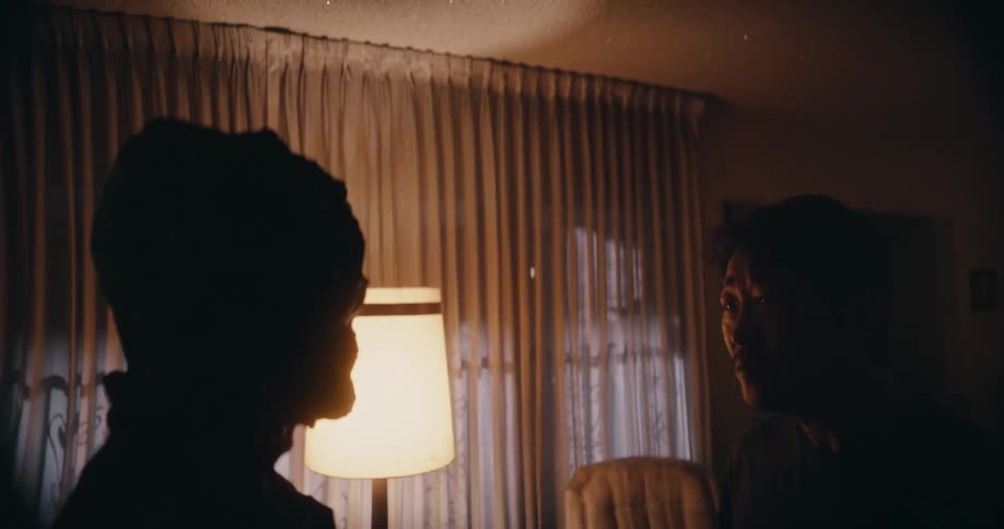 A man is standing in his living room with fear on his face as a creature with a strange morphing face made of a dark organic substance stands in front of him