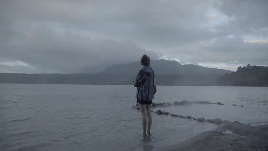 A woman stands ankle deep in water at a misty shoreline