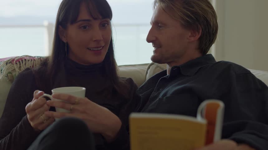 A man and woman sitting on a couch together while the man holds a book and the woman holds a coffee mug