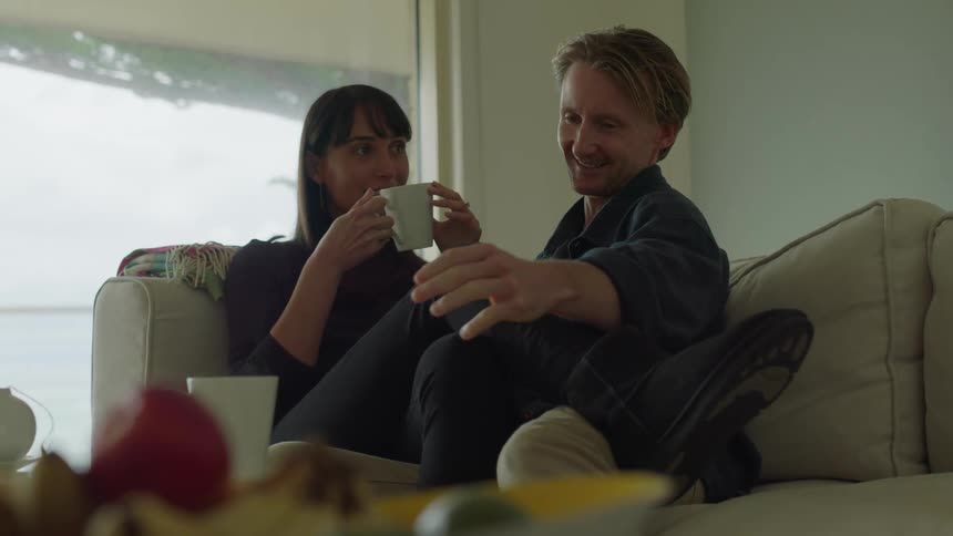 A man and woman sitting together on a couch as they kiss and both hold a white mug