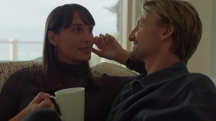 Man in blue shirt and woman with black sweater kiss each other and laugh while holding cup in the living room