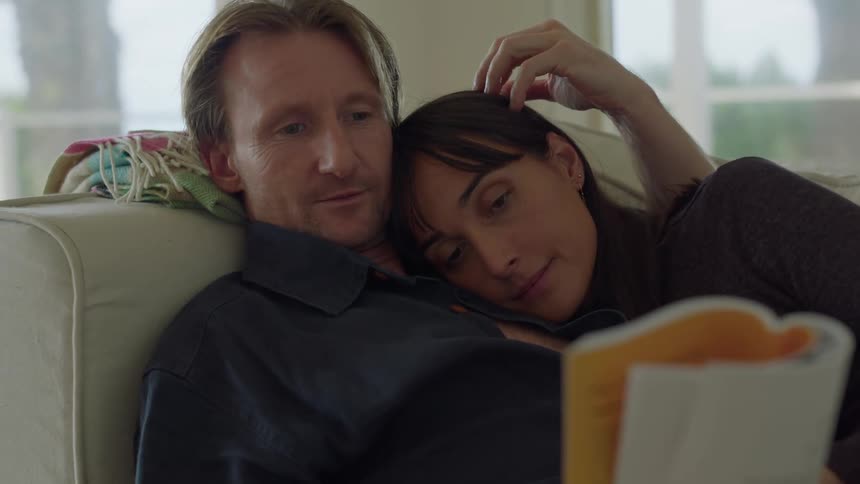 A man and woman cuddle together on a couch while the man reads