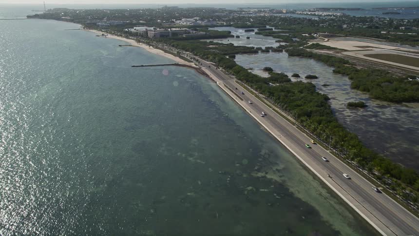 A breathtaking aerial view of South Roosevelt Boulevard in Key West Florida as cars drive over the coastal road and a suburban neighborhood seems to emerge on the horizon