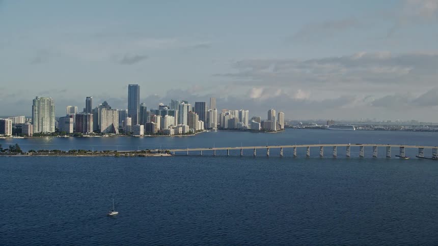 Aerial footage captures the stunning beauty of Miami’s skyline featuring the Rickenbacker Causeway bridge and skyscrapers amidst a partially cloudy sky during sunrise