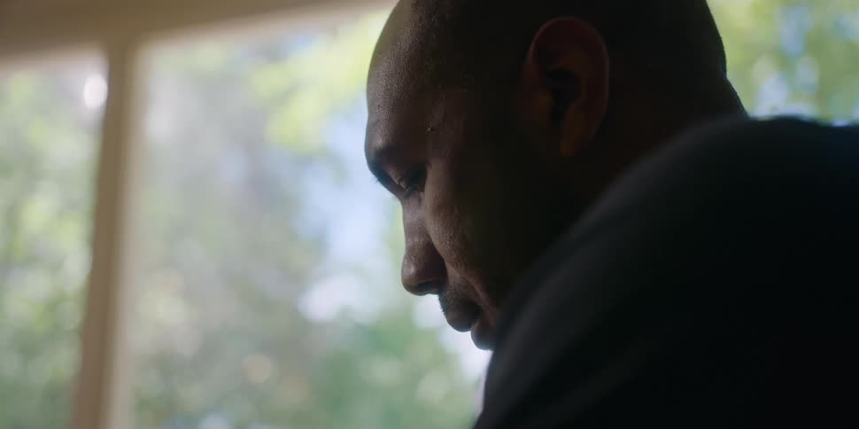 A sad looking man sits in front of a large window at home and looks up