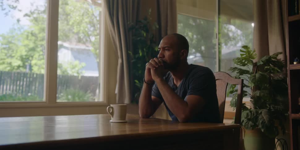 A black man sits at a table pensive with a mug in front of him
