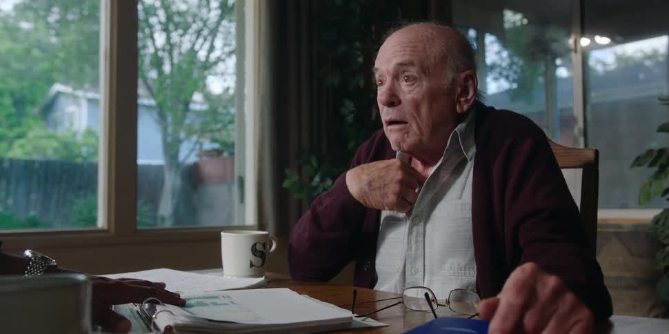 An elderly man sitting at a table frowns and points to himself as he talks with someone offscreen