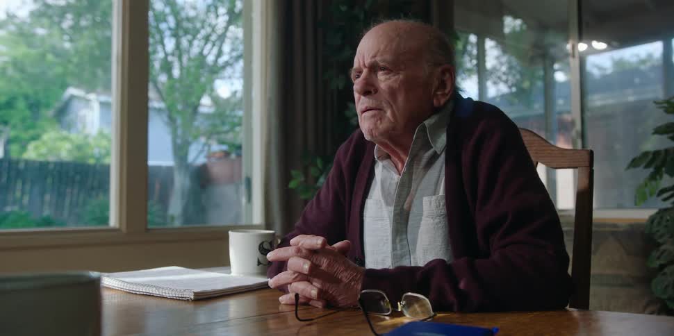An elderly man is sitting down at a desk talking and he reaches out and shakes another mans hand