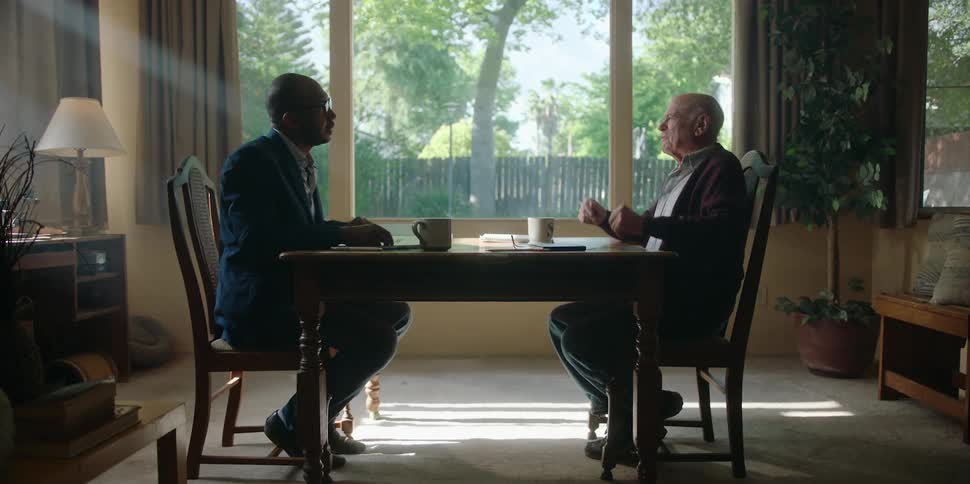 An elderly man sits at a table at home and talks with a business man across from him