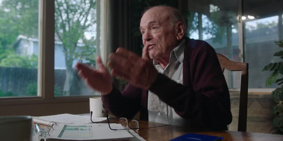 An elderly man sits at home and laughs and looks at documents as a man points at them