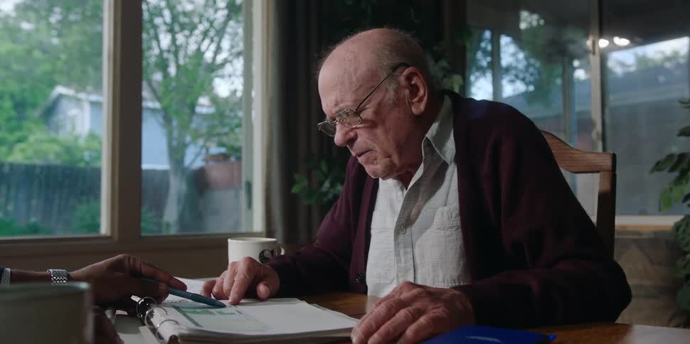 An elderly man at home looks confused as he looks over documents with another man