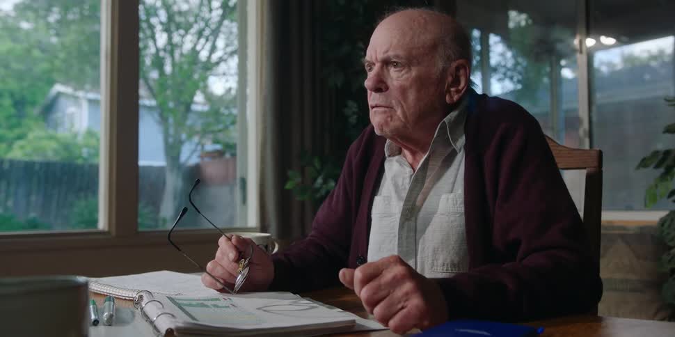 A man holds his glasses while talking about papers strewn across a table