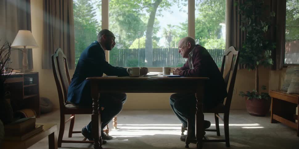 Two men are sitting at a table talking as the elderly man puts on his glasses and the other man points to documents