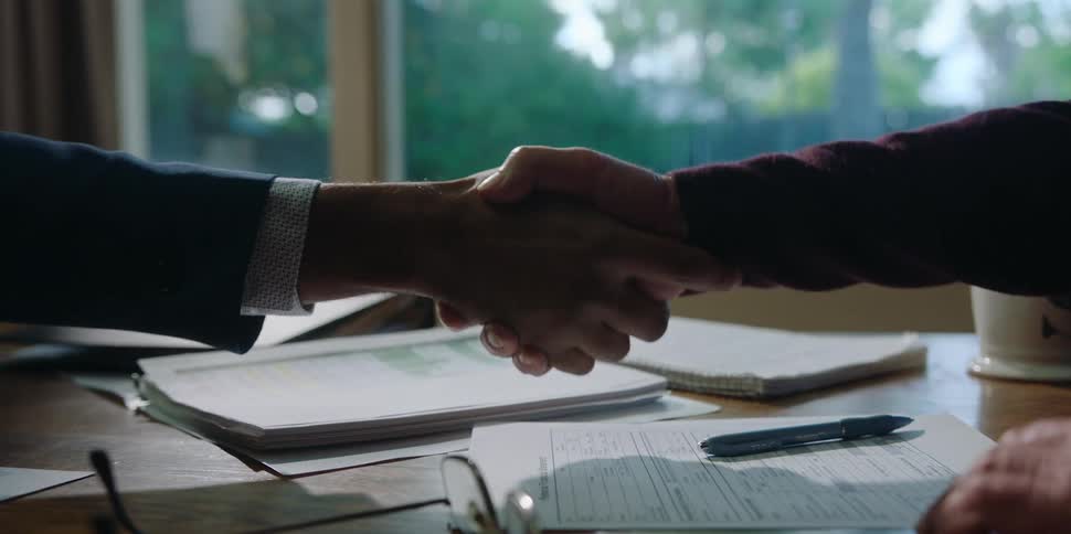 An elderly man at home signs a document and reaches across a table to shake hands with a man