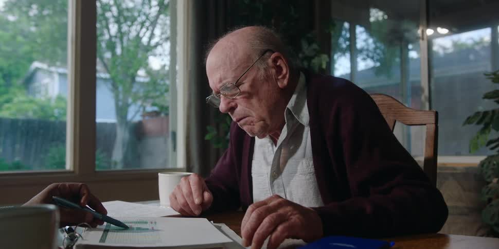 An old man is talking and puts on his glasses when someone shows him a page from a dossier that surprises him then he takes off his glasses and puts them on the table in front of him