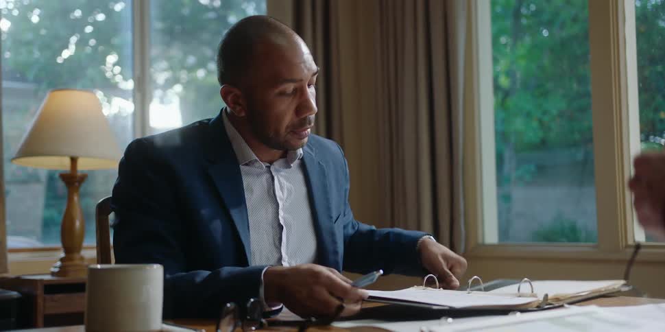A man is sitting at a desk as he takes off his glasses and opens a notebook and takes out a document and looks over to speak to the person across the desk as he holds a pen in his hand