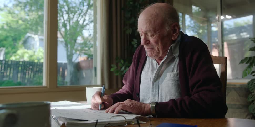 An elderly man is sitting down at a desk holding a pen as another man points to the document and the man signs it