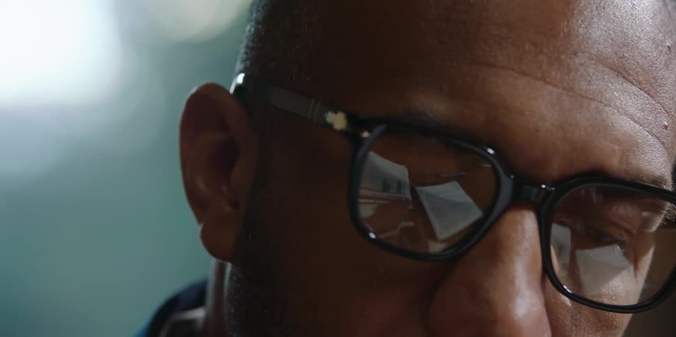 A man wearing glasses is looking down as the reflection of documents show on the lenses and he looks up