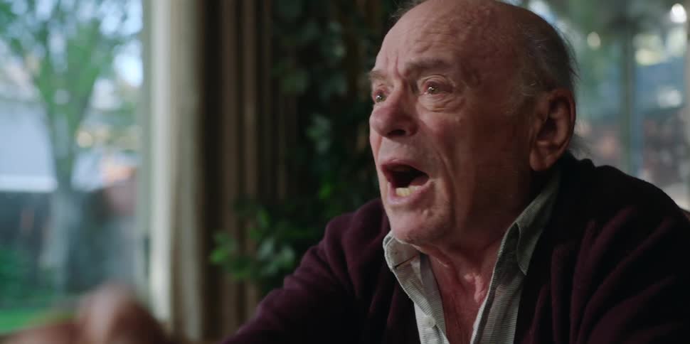 An elderly man is sitting down moving his hands and becoming emotional as he speaks