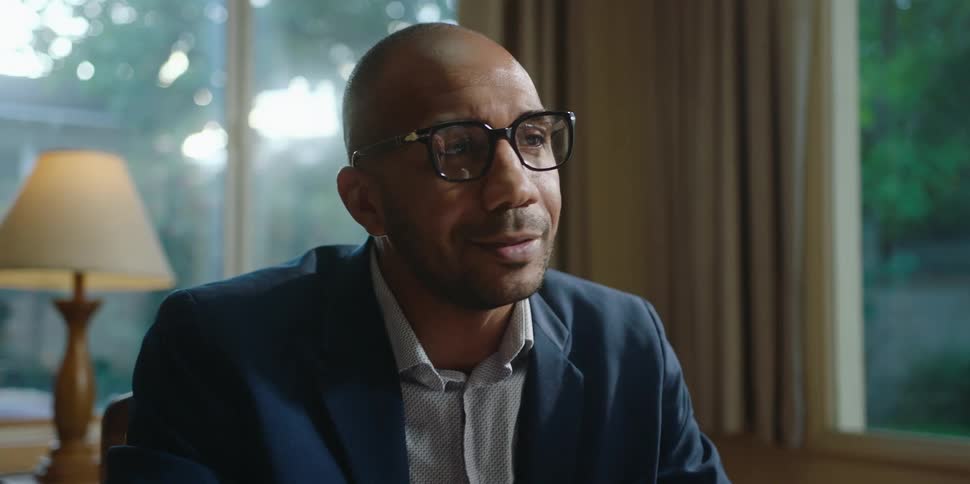 A man wearing glasses is smiling and nodding his head as he speaks to another person