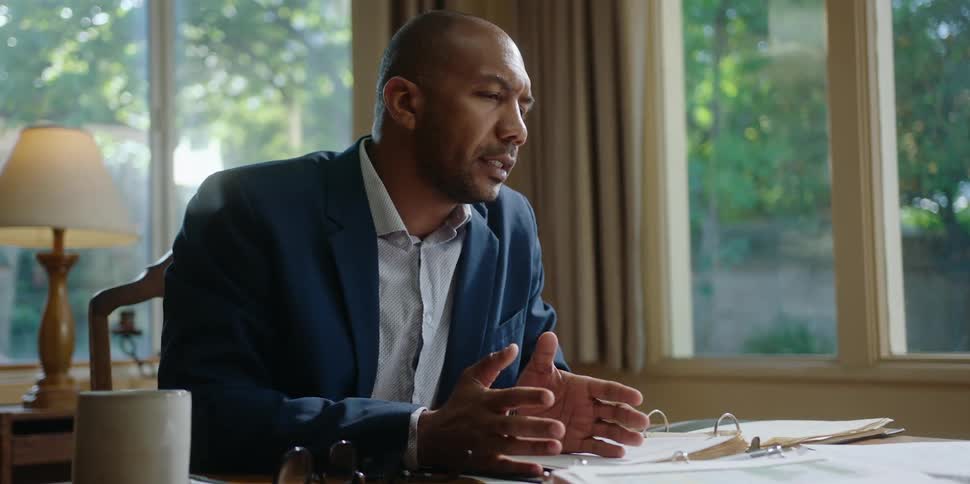 A man is sitting at a desk with an open notebook and moving his hands as he speaks to another person