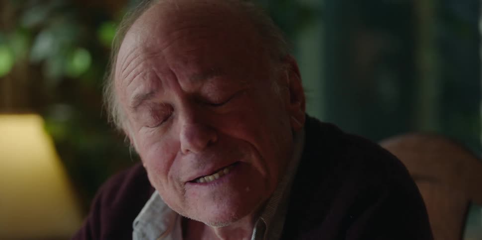 An elderly man at home talks and smiles as he lifts his hands up next to his head