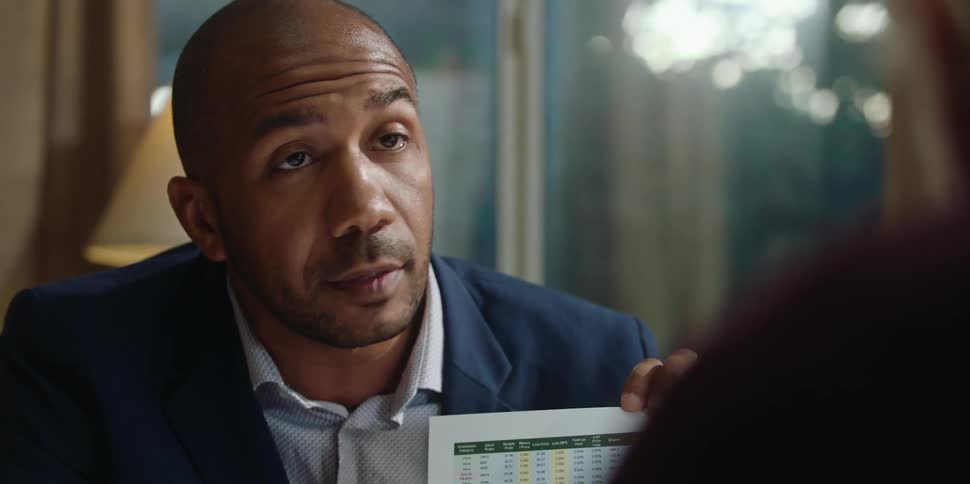A man is sitting down talking to another man and holding up a document and pointing to a chart as he speaks