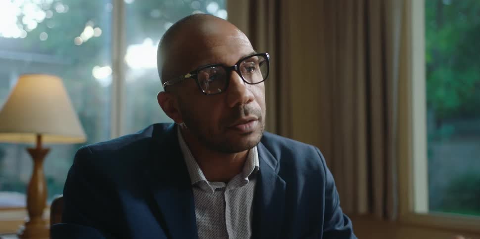 A man is sitting indoors and wearing glasses
