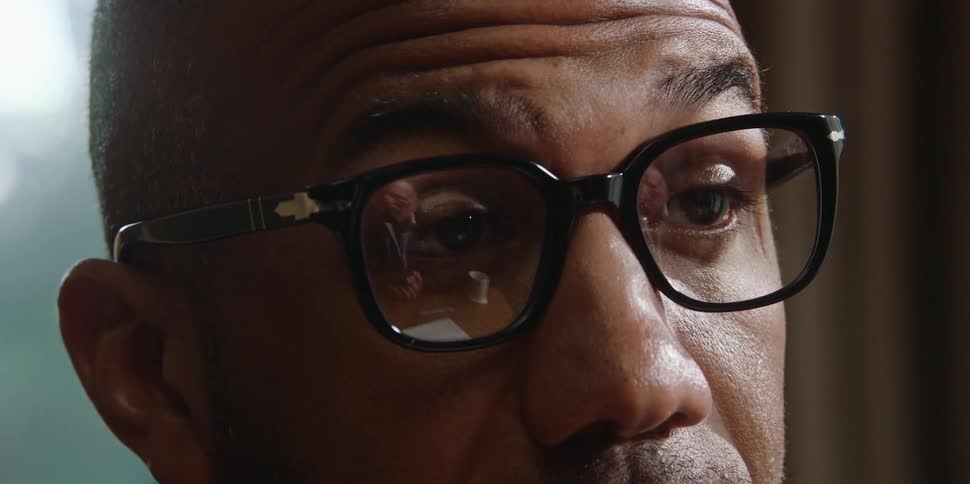 A black man with glasses is talking to an elderly white man reflected in the glasses