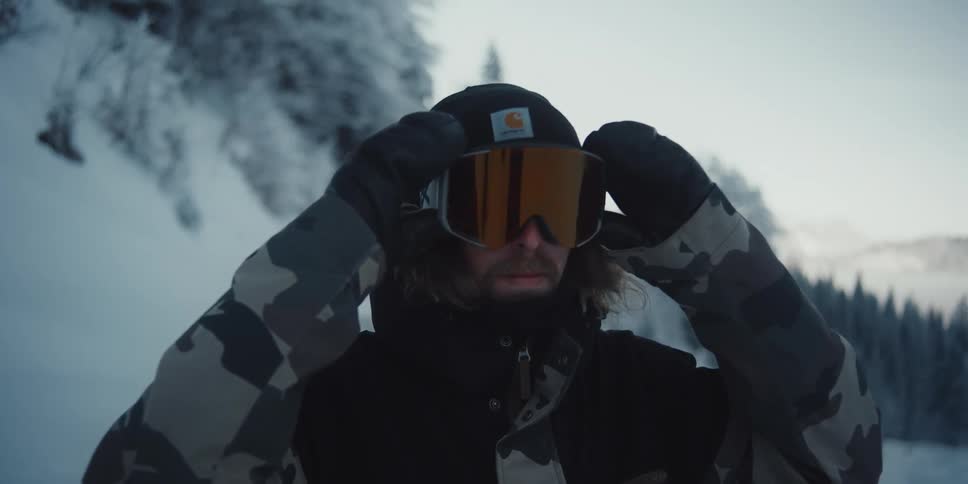 A young mans on a snowy mountain range lowers goggles over his eyes