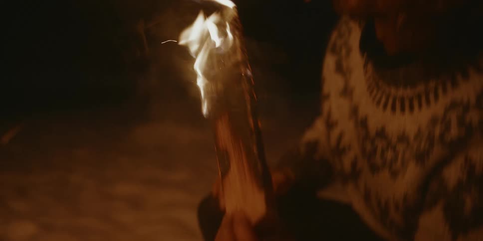A man takes a piece of burning wood from a campfire and lights his cigarette with the flame