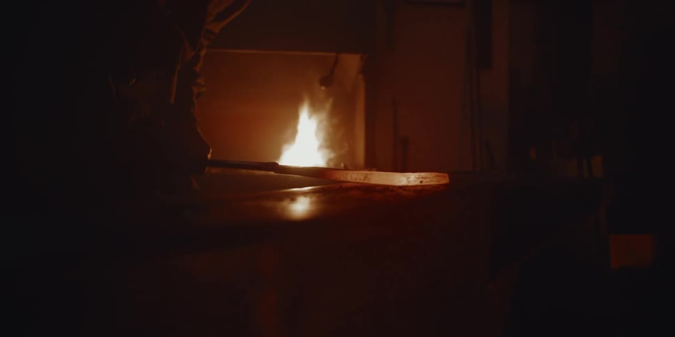 Blacksmith hammers glowing metal wedge in smithy near flame